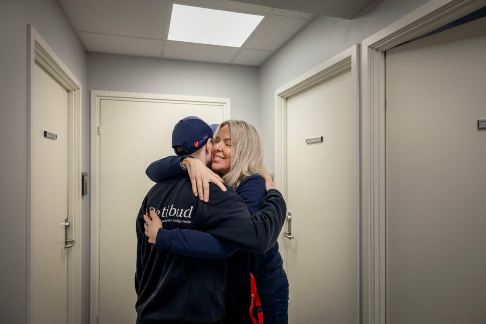 Foto: Kvinne med kort, mørkt hår og caps klemmer en blond dame med langt hår i en korridor.