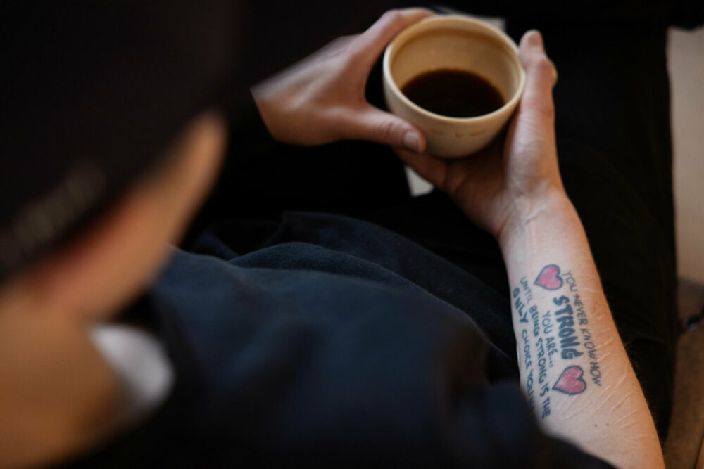 Foto: To kvinnehender holder i en kaffekopp. På den ene armen er det en tatovering med mye tekst og to røde hjerter.