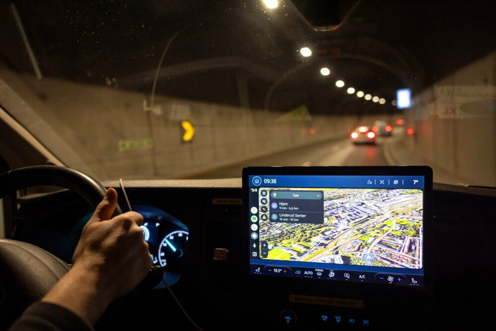 Foto: En hånd på rattet og en skjerm som lyser opp med et kart. Bilen kjører i en tunnel, så det er mørkt.