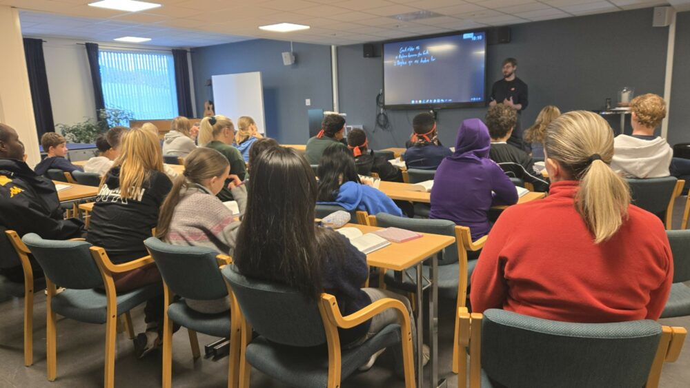 Ca. 40 konfirmanter, som vi ser bakfra, sitter på stoler i et slags auditorium. En voksen person står foran og underviser.