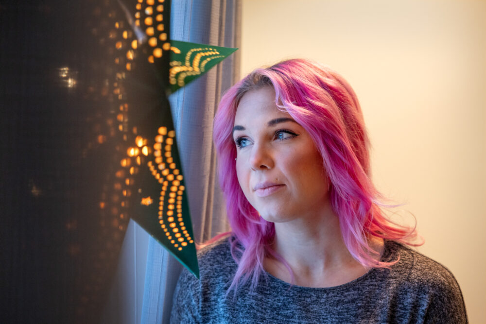 Kvinne med rosa hår som ser drømmende ut av vinduet. Der henger det et grønn, stor papirstjerne med lys i.