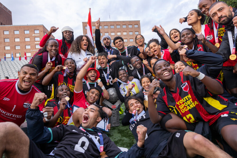 Foto: Fire gutter av afrikansk opprinnelse i båt innover mot rådhuskaien i Oslo. De står med ryggen til i grønne drakter. Alle har på seg svarte hettegensere der det står "Homeless World Cup, street soccer, Oslo Norway 2017. Frelsesarmeens gatefoball.