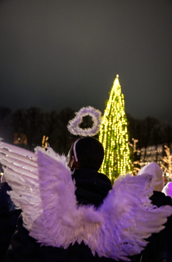 Foto: Barn som synger sett bakfra med hvite enklevinger på. Langt der borte ser vi en diger, tent julegran.