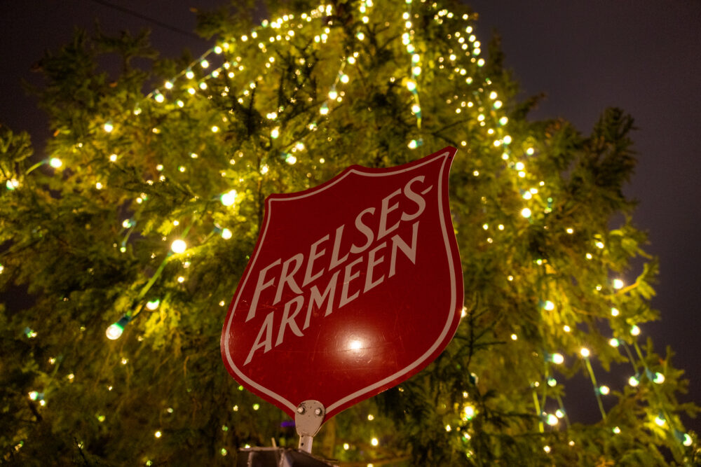 Foto: Nærbilde av skiltet i toppen av julegrytestativet, som da er Frelsesarmeens røde logo, opp mot tusenvis av lys i et digert utendørs juletre.