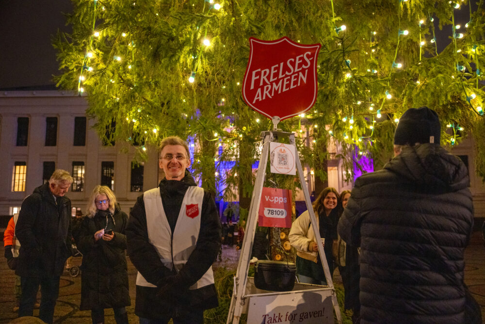Foto: Mann fra Frelsesarmeen i hvit refleksvest med logo på. Han står ved julegryta og rundt ham er det vinterkledde folk og et opplyst juletre.