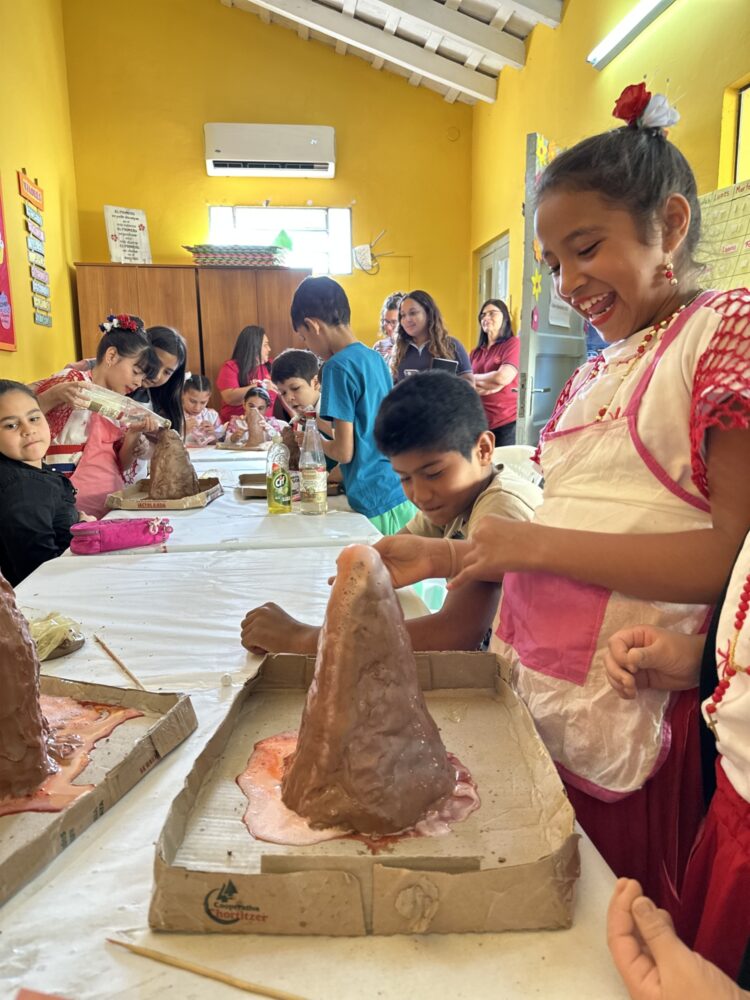 Foto: Jente står og ler mens hun lager en vulkan av leire eller liknende på skolen. Mange barn i bakgrunnen.