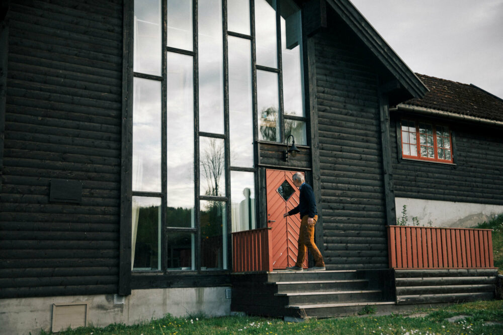 Foto: Mann som åpner en rød dør inn til et stort, brunt bygg med en hel vegg av vinduer. Bygget ser nesten ut som en moderne kirke, men med gammel inngangsdør. Den har firkantet vindu til å titte inn gjennom der vinduet er snudd 45 grader slik at spissene kommer opp, ned og ut til kantene av døra.