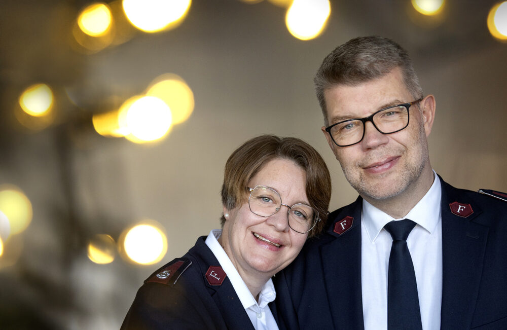 Foto: Kvinne og mann i 50-årene lener seg mot hverandre. I bakgrunnen er det mange lys. Begge bruker briller og har på seg uniforma fra Frelsesarmeen.