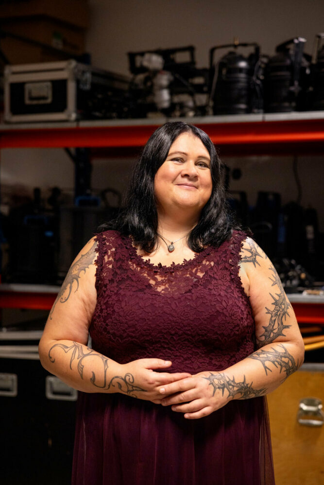 Foto: Kvinne med mørkt hår i burgunder blondekjole uten ermer. Tatoveringer slynger seg oppover på begge sider.