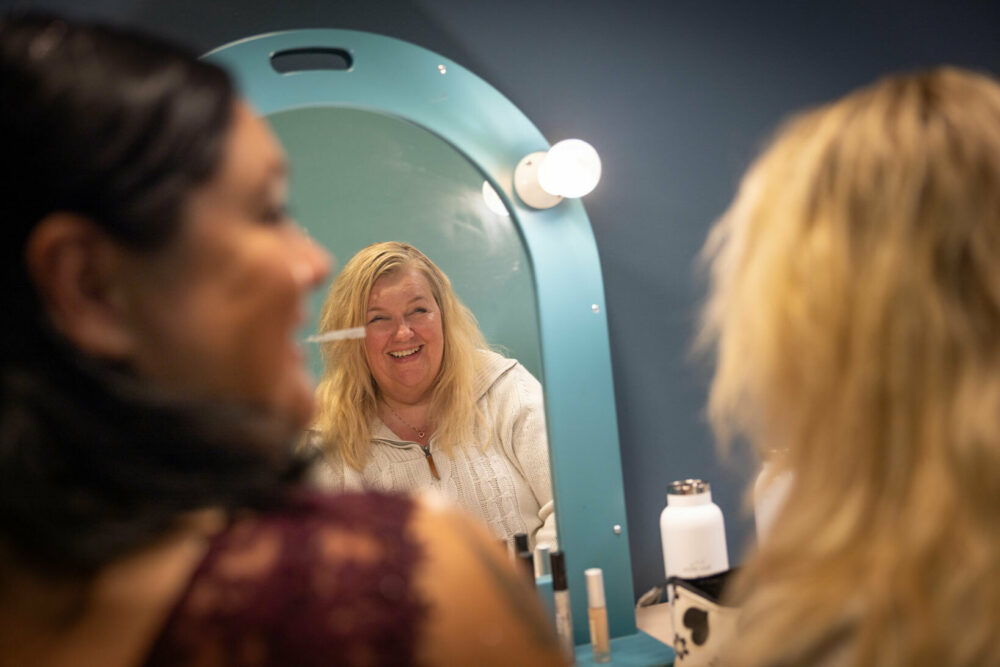 Foto: To kvinner, en mørk og en blond sitter foran speilet. Hun blonde smiler godt mens hun mørke ser på henne.