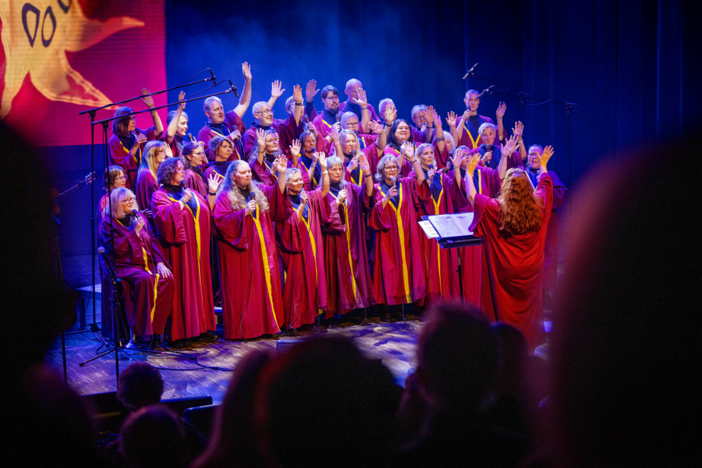 Foto: Gospelkor som synger fra en scene og vinker. De har på seg røde kapper. Med ryggen til publikum står en dirigent med langt, rødt hår.