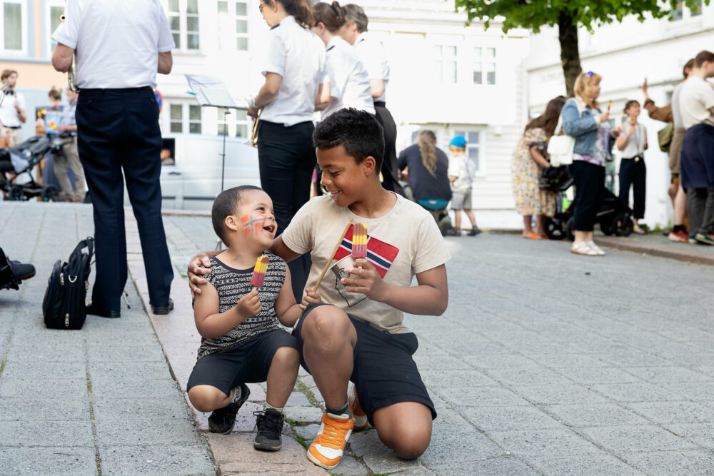 Foto: Av to barn med mørkt hår og krøller som sitter på huk. Se spiser is og smiler mot hverandre. De er dekorerte med norske flagg, den eldste på T-skjorta og den yngste på kinnet.