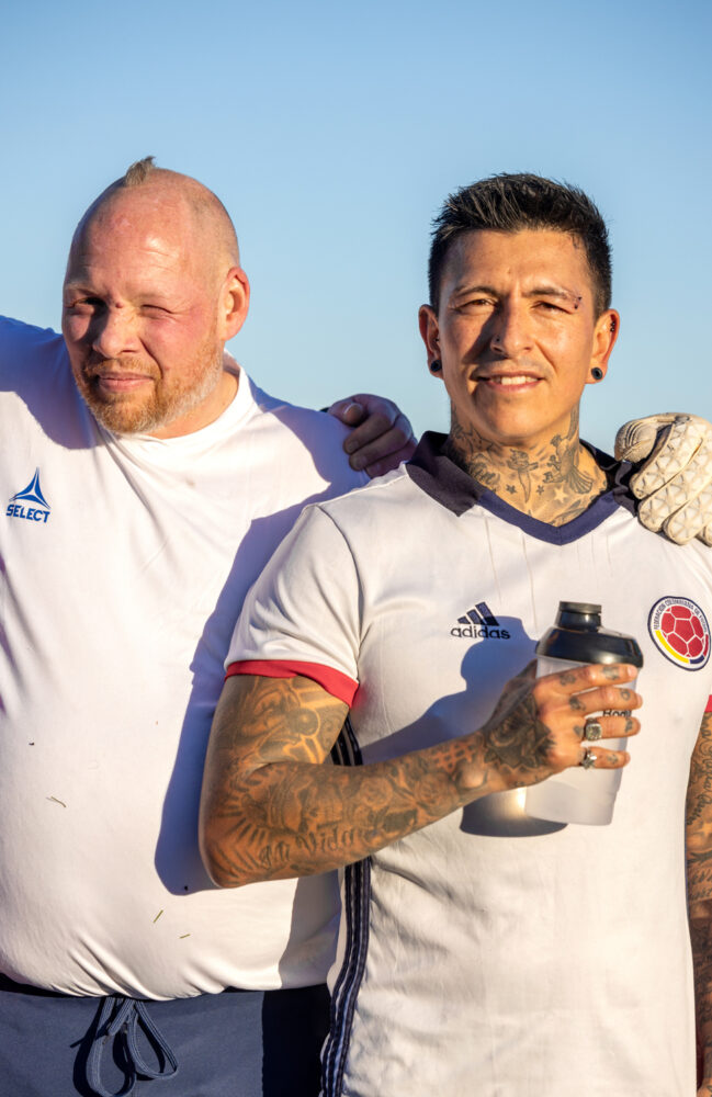 Foto: To menn står tett ved siden av hverandre på en fotballbane. Den ene blond i håret, den andre svart. Begge har på seg hvite drakter og myser mot sola. Han til høyre holder en vannflaske.