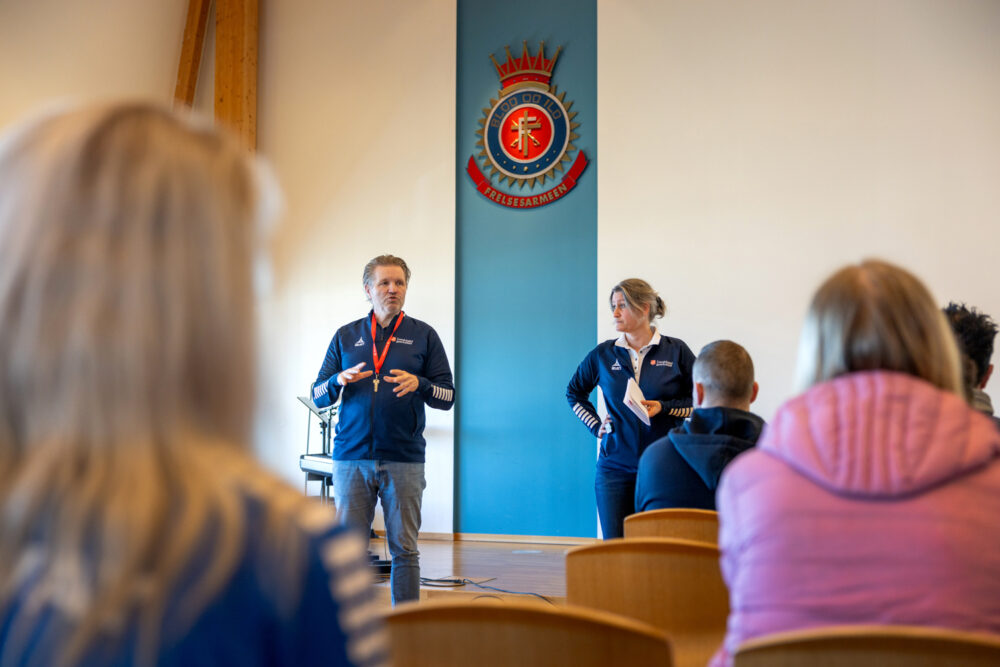 Foto: Mann og kvinne i blå treningsjakker står og snakker foran en gruppe kvinner og menn. På veggen bak henger Blod og ild – et skilt i Frelsesarmeen.