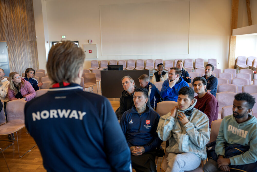 Foto: Mann med ryggen til, der det står NORWAY, snakker til en gruppe kvinner og menn som sitter i en sal.