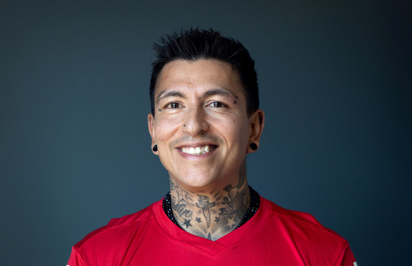 Foto: Mann med gyllen hud og kort, svart hår smiler i en rød fotballdrakt med norsk flagg på. Han har mange tatoveringer oppover halsen.