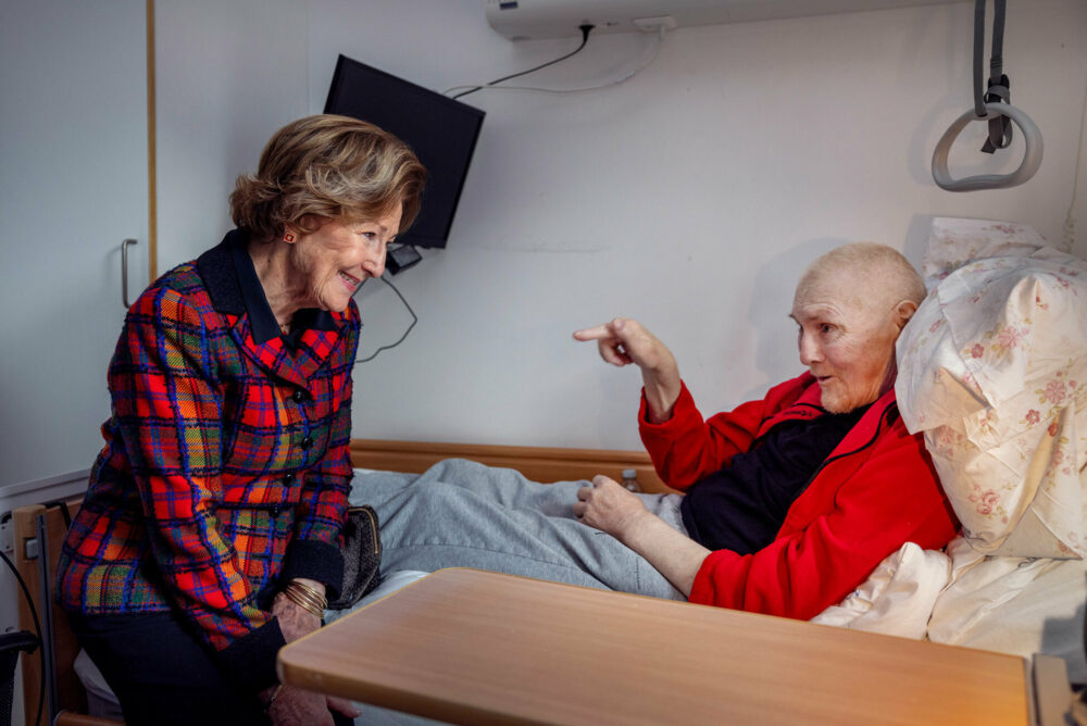 Foto: En kvinne i 80-årene med håret pent frisert sitter på sengekanten til en mann uten hår i 70-årene. Han ligger i en sykeseng med rød flisjakke på seg. Han peker mot damen.