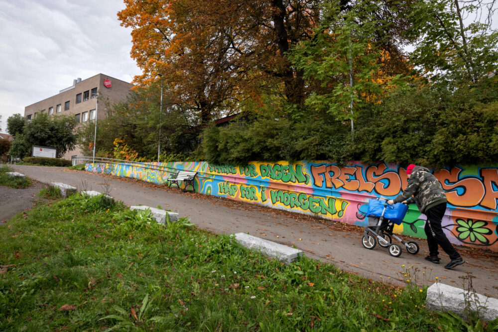 Foto: Mann i armyjakke og rullator går oppover en bratt bakke med fargerik grafitti på muren ved siden av veien. Mursteinsbygget på toppen har en stor, rød logo fra Frelsesarmeen på veggen.