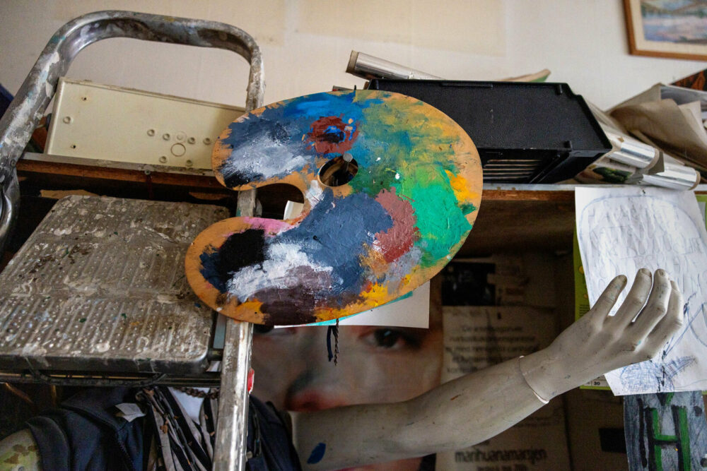 Foto: En hånd til en manequin stikker ut fra en bokhylle med mye rot. En gardintrapp står lent opp mot den og en buet plate med ulike fargeklatter henger på en spiker.