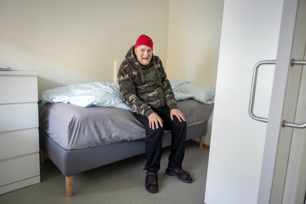 Foto: Den eldre mannen i armyjakke sitter på en oppredd seng. Det er rent sengetøy på og sengen og den hvite kommoden ved siden av er begge nye.