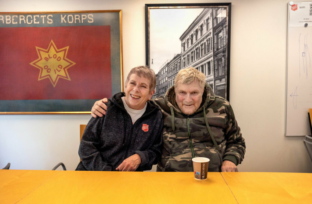 Foto: Den eldre mannen i miltærjakken sitter ved siden av en kvinne i 60-årene. Han har armen godt om hennes skulder. Begge smiler godt. På veggen bak dem henger et gammelt foto av en bygning. Ved siden av på veggen henger et innrammet, rødt flagg fra Frelsesarmeen med en gul stjerne på der det står "blod og ild".