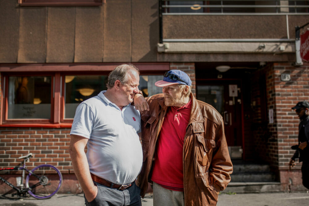 Foto: To menn står tett ved siden av hverandre og snakker ute på gata foran en rød murbygning, som er fyrlyset. Den ene er i 50-årene og den andre i 60-årene.