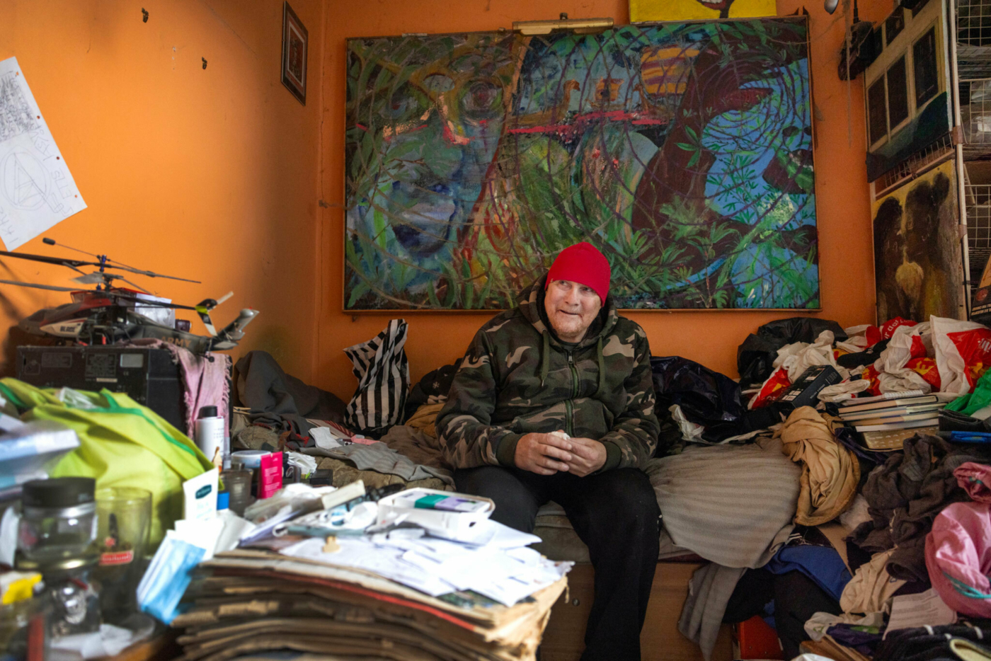 Foto: Mann med rød lue og jobbejakke i militærmønster sitter pp sengekanten. Over senga henger et stort maleri. I enden av senga og på gulvet står stabler av bøker, aviser og plastposer med ting i.