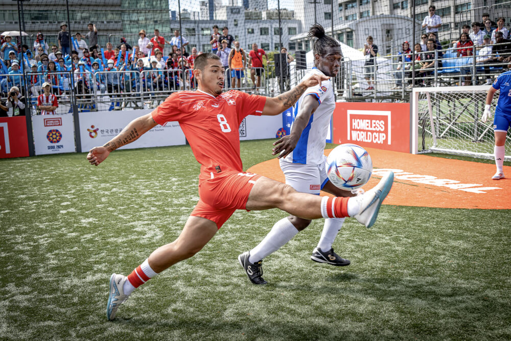 Foto: To mannlige fotballspillere, en i rød drakt og en i hvit/blå i en ballduell like foran et mål. På tribunen bak sitter det mange mennesker og heier. Bildet er fra Seoul i 2024.