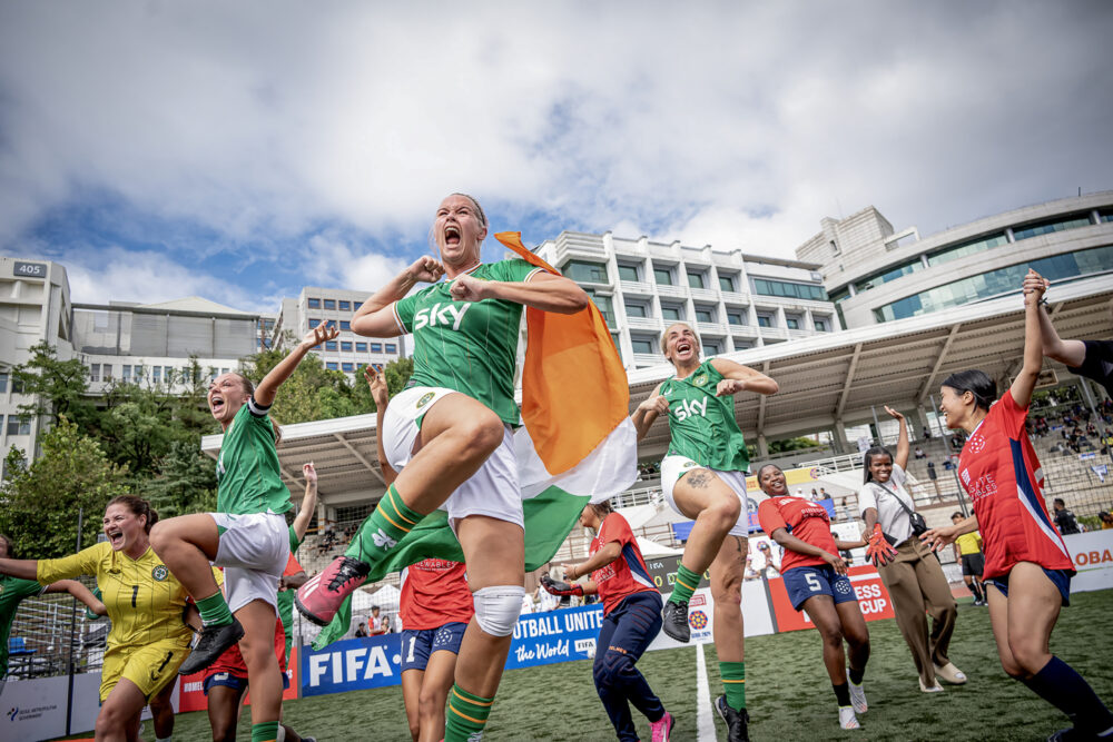 Foto: Kvinnelige fotballspillere i grønne/hvite og røde/blå drakter hopper og danser på en liten fotballbane i Seoul i 2024.