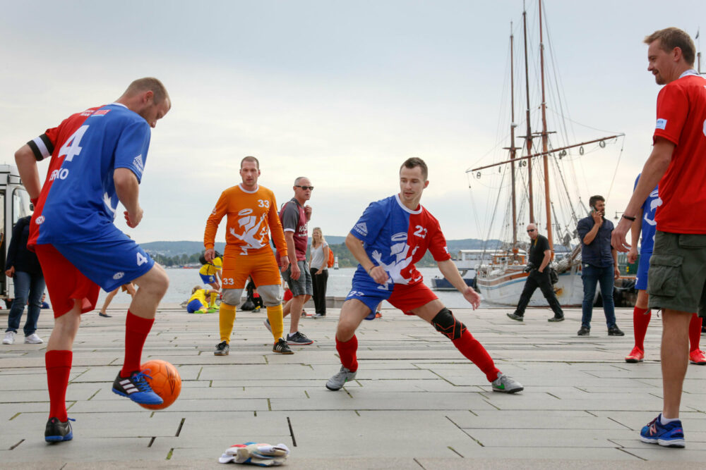 Foto: Menn i litt ulike fotballdrakter varmer opp rett på steinhellene på rådhusebrygga i Oslo. Like nedenfor ligger ei seilskute til kai.