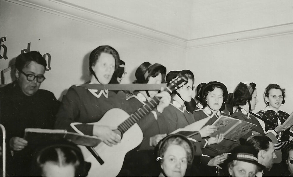 Foto: Gammelt svart-hvitt-bilde av kvinner og menn i uniformer fra Frelsesarmeen. De synger og mange spiller også gitarer.