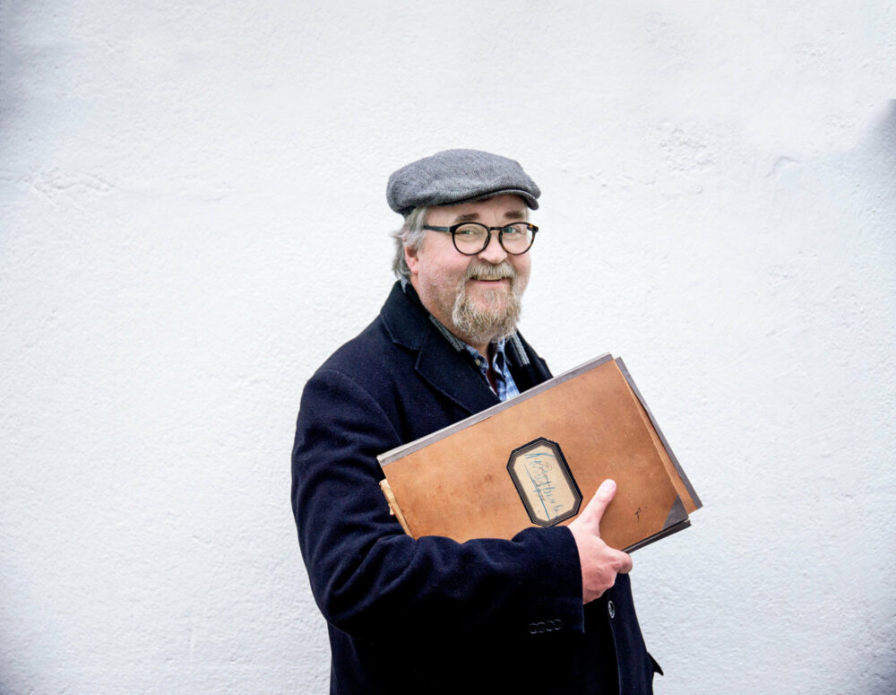 Foto: Mann med sixpence på hodet og runde, brune briller. Han er i slutten av 50-årene og holder flere gamle mapper i skinn under høyre armen.