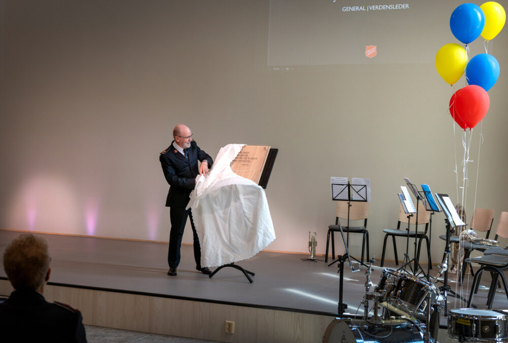 En middelaldrende mann med mørk uniform på en scene. Han drar bort et klede som dekker en plakett.