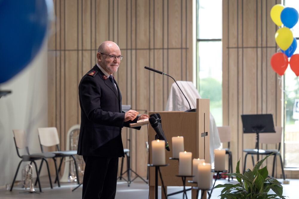 En smilende, skallet, middelaldrende mann med briller og sort frelsesarméuniform foran en mikrofon og en talerstol på podiet i et stort forsamlingslokale. Veggene bak har store vinduer og lyst treverk.