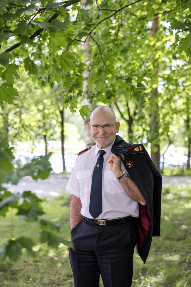 Portrettfoto av en smilende, skallet, middelaldrende mann med briller, hvit skjorte og sort frelsesarméuniformsjakke over skulderen. Bak ham er det grønne trær og gress.