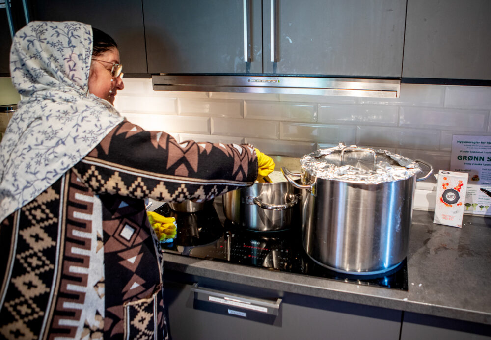 Foto: En kvinne med sjal over håret og brun og svart mønstrete kjole sees fra siden, hun står ved komfyren og rører i to fulle gryter.