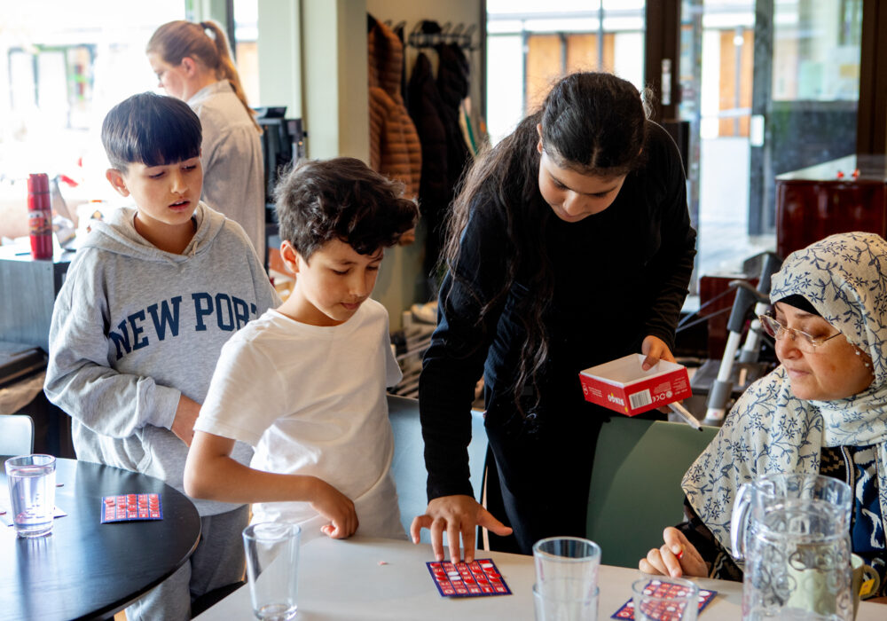Foto: Rand står i midten av bildet med svart langt hår og svarte klær. Hun bøyer seg over et bord og sjekker et bingobrett. To gutter står til venstre og følger med, til høyre sitter en kvinne med sjal over håret og ser på.