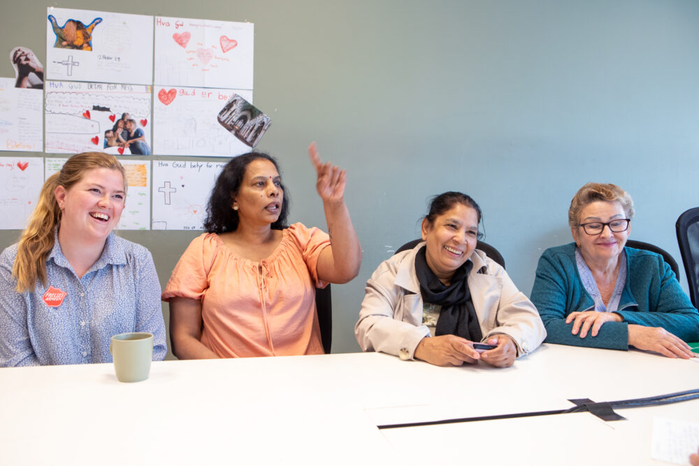 Foto: Åse Kari i blå skjorte med Frelsesarme-skjold sitter ved et bord ved siden av tre kvinner. Alle smiler og ser mot siden.