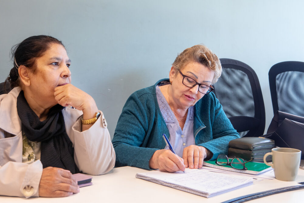 Foto: Bildet viser to kvinner som sitter ved et bord. En kvinne med mørkt hår og beige jakke sitter og ser mot siden, ved siden av en kvinne med lyst, kort hår og turkis strikkejakke som skriver på et ark.