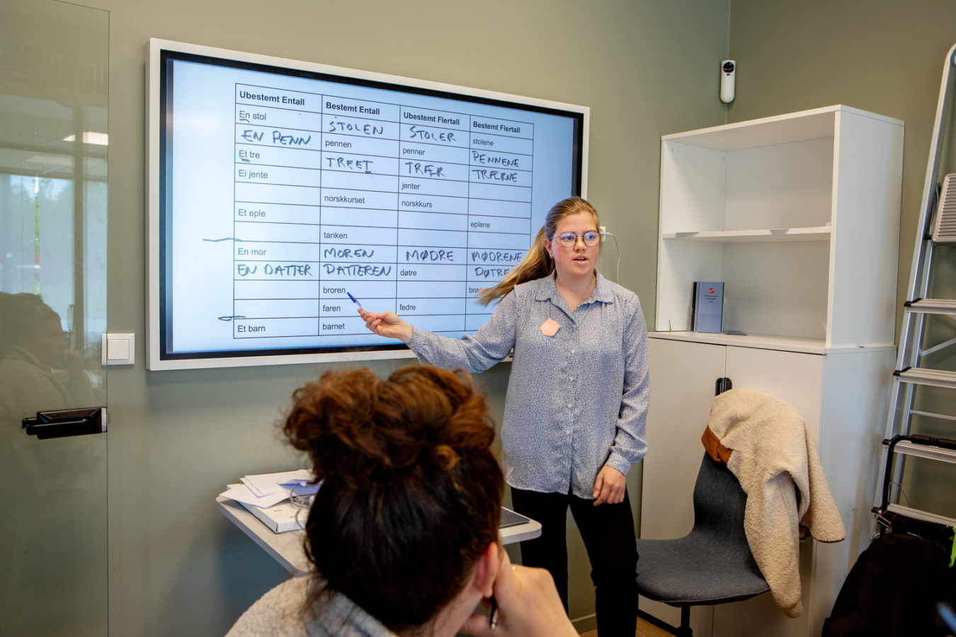 Åse Kari, med langt lyst hår og blå skjorte med frelsesarme-skjold, står og peker på en skjerm der det er skrevet bøyninger av norske ord. Bakhodet til en av kursdeltakerne sees i bakgrunnen.