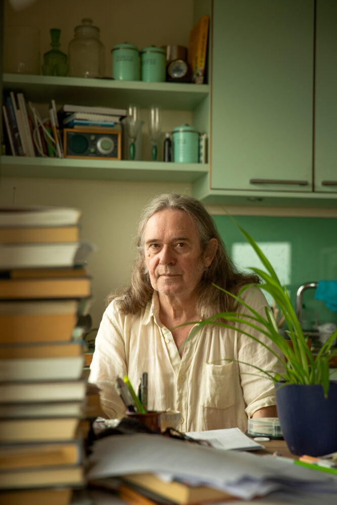 Foto: Mann med halvlangt hår og beige skjorte sitter ved et skrivebord, ved siden på bordet er det en stabel med bøker.