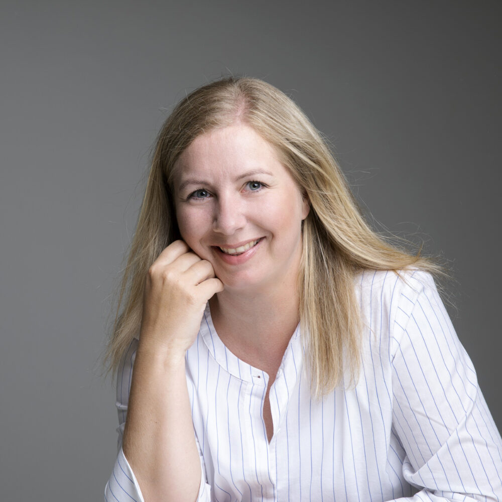 Janette Røseth sitter og smiler mot kamera. Hun lener seg på den ene armen, hun har på seg lys stripete skjorte og bakgrunnen er grå.