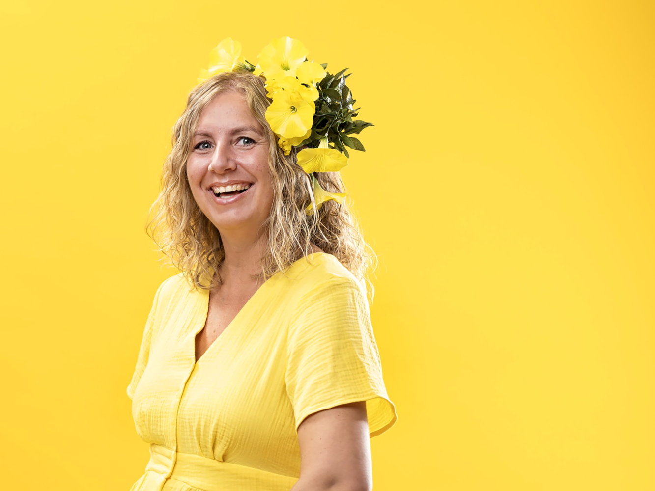 Bildet viser Janette Røseth som smiler mot kamera. Hun har lyst, langt hår, gul kjole, gule blomster i håret og bakgrunnen er gul.