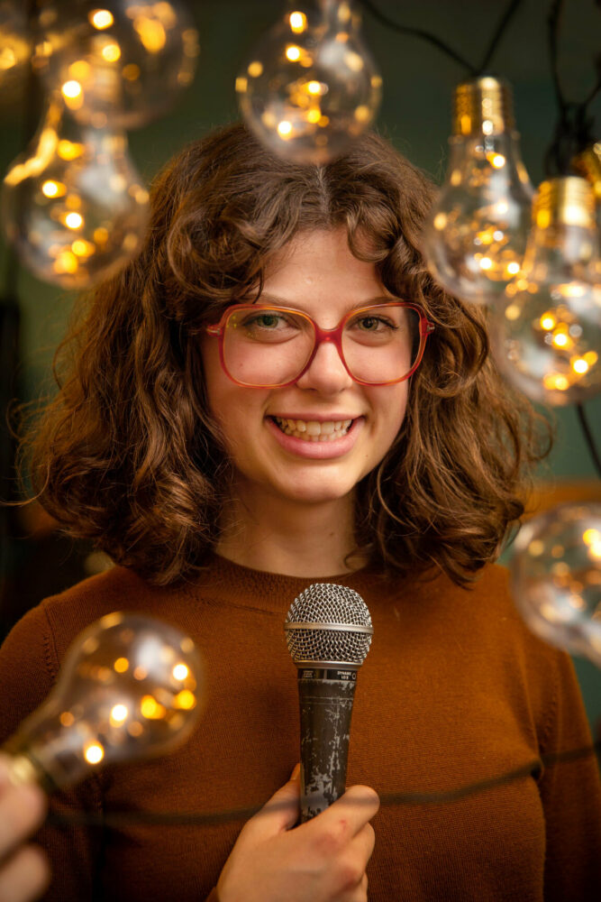 Foto: Ung kvinne i brun genser med store briller og mørke krøller holder en mikrofon og ser i kamera. Rundt henne er det en masse store lyspærer med varmt lys i.