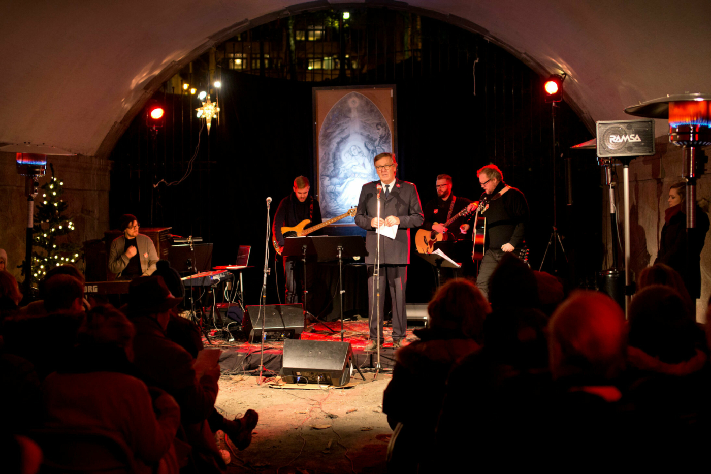 Foto: Mann i uniform fra Frelsesarmeen står og snakker i en mikrofon under en bro. Det er mørkt ute, men her er det pyntet med lys. Rundt ham står og sitter bandmedlemmer med instrumenter.