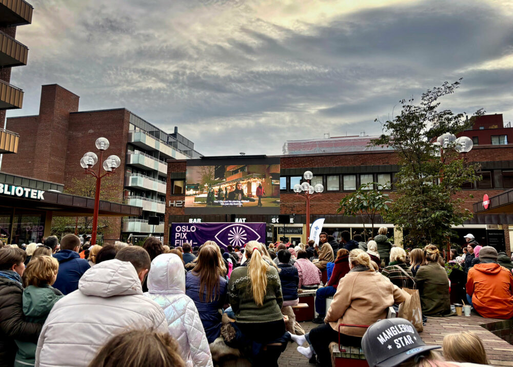 Foto: Masse mennesker sitter i boblejakker med ryggen til kamera. De er på et torg og ser på en film som vises på veggen til et av byggene i rød murstein.