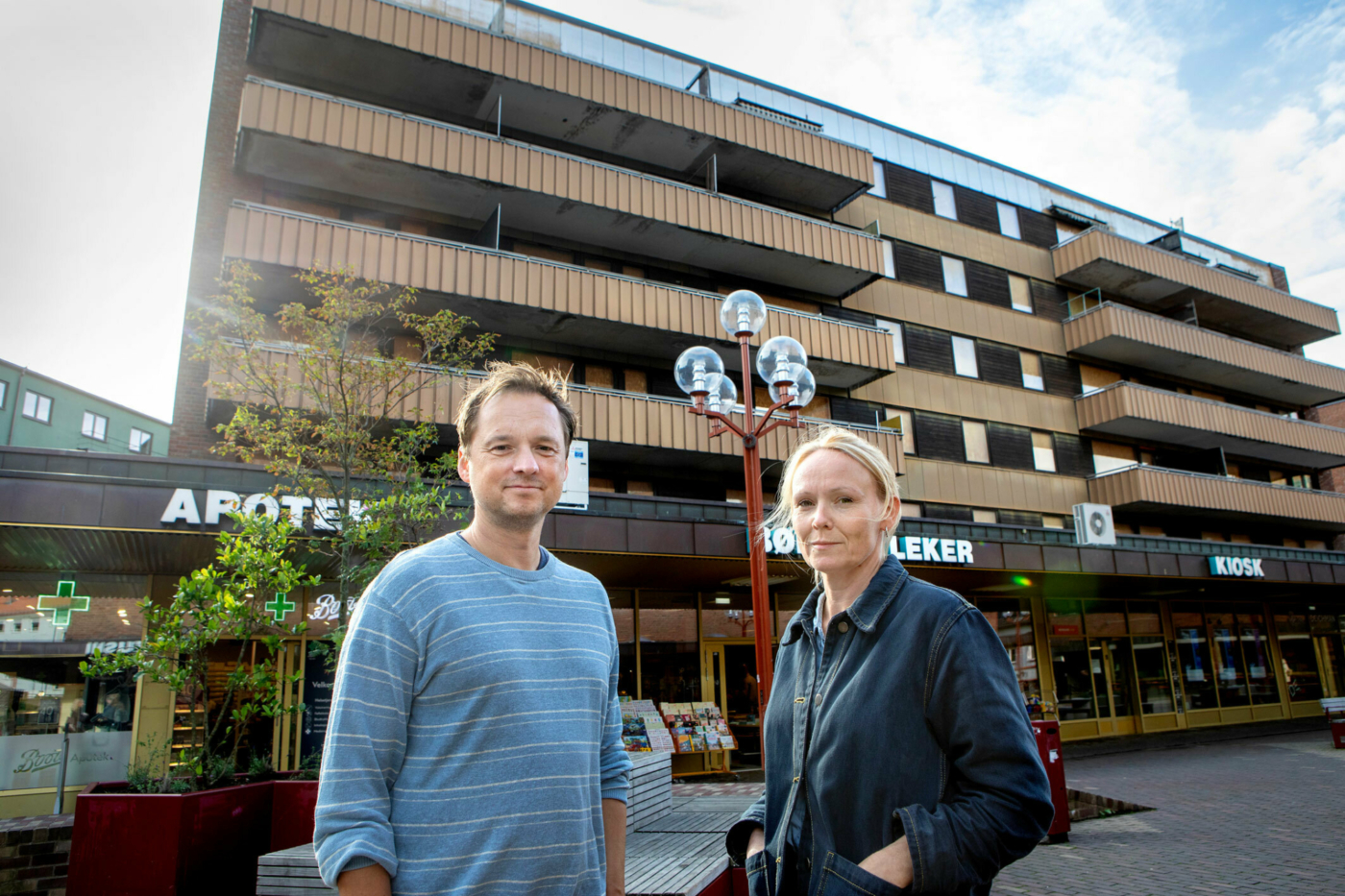 Foto: Utenfor en sliten blokk i Oslo står en mann i 40-årene med mørkeblondt hår i en mellomblå genser med striper. Ved siden av ham står en jevngammel, blond kvinne i dongerijakke.