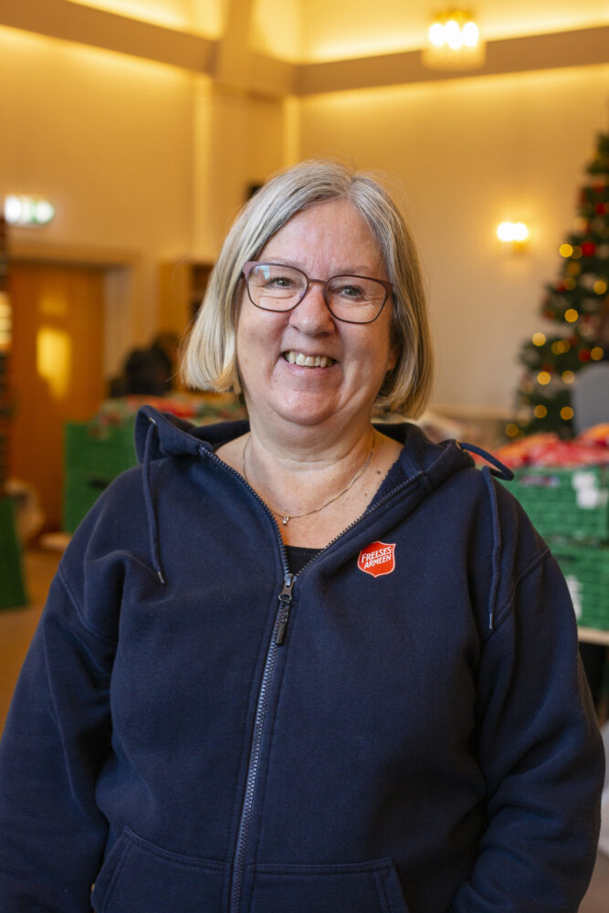 Foto: Kvinne med grått hår og briller smiler. Hun har på seg blå flisjakke og rød logo fra Frelsesarmeen. Står i et stort rom med "julerigging" i bakgrunnen.