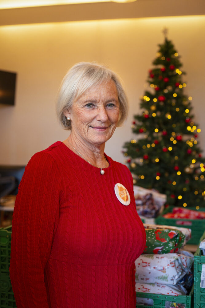 Foto: Pensjonert kvinne i rød genser med en pin fra Frelsesarmeen på brystet smiler til fotografen. I bakgrunnen er det både juletre og gaver oppstilt.