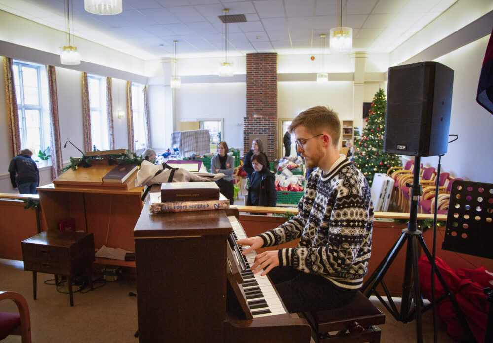 Foto: Ung mann med briller og skjegg spiller piano i et kirkerom. I salen er det flere frivillige og stabler med gaver.